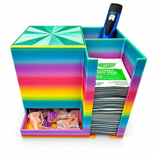 Rainbow insulin pen organizer with a green geometric lid, packaged needles, a blue injection pen and stacked alcohol prep pads on white. Supplies not included.